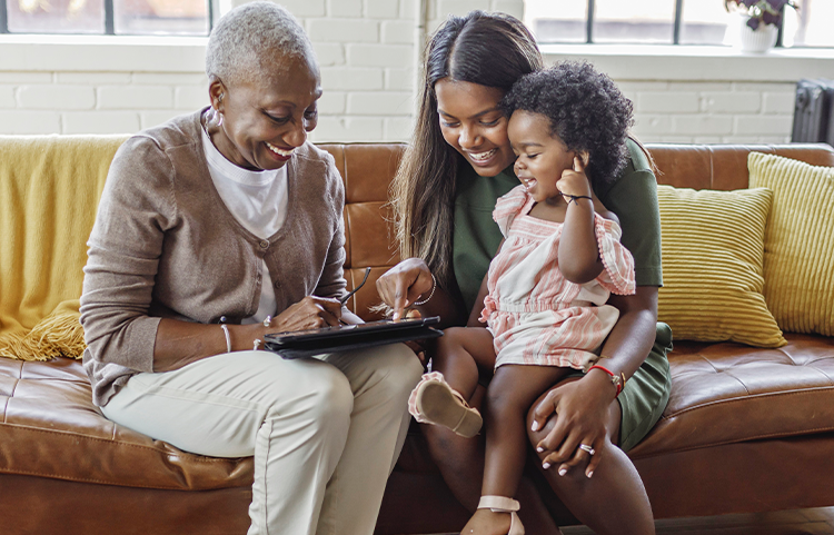 Three generations (a young girl, her mother and grandmother) smile on a couch while looking at an electronic device.